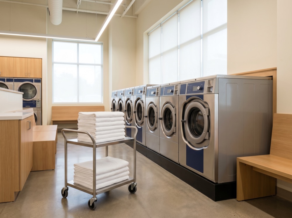Row of large commercial washers and dryers with a cart of folded linens